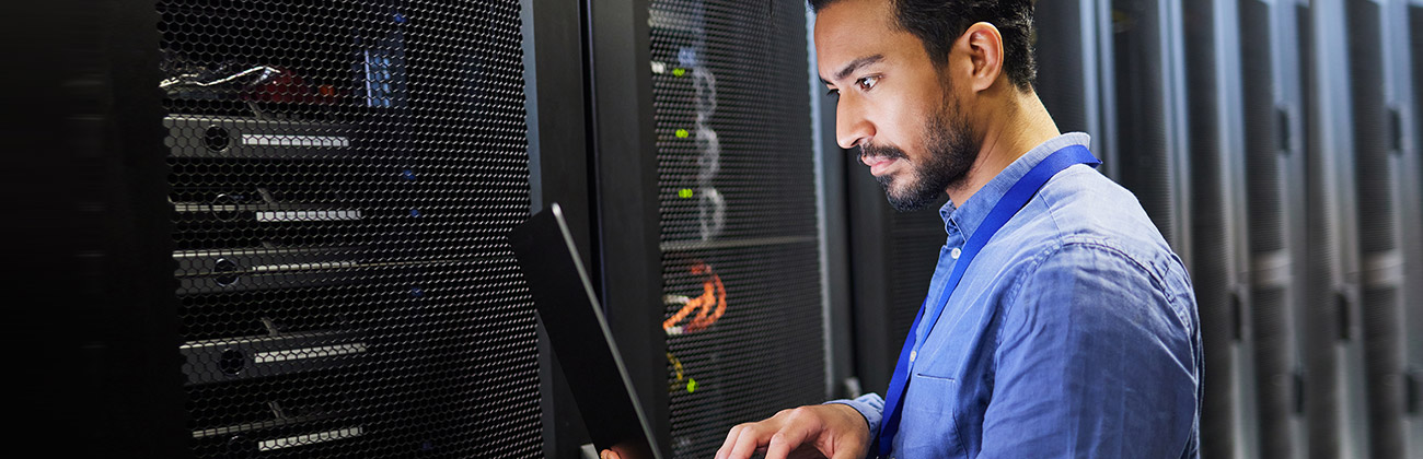 Man keys into laptop in a data center surrounded by stacks of servers.