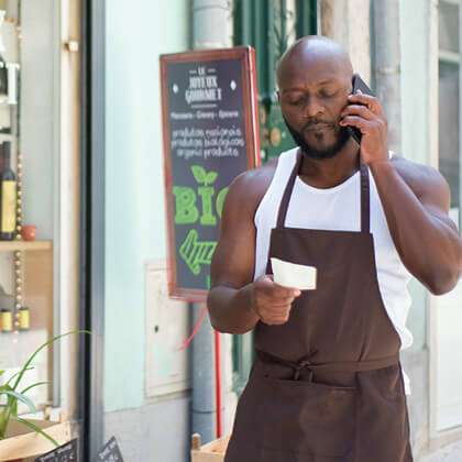 Dark-skinned man in a trendy grocer apron uses mobile phone outside a boutique gourmet shop