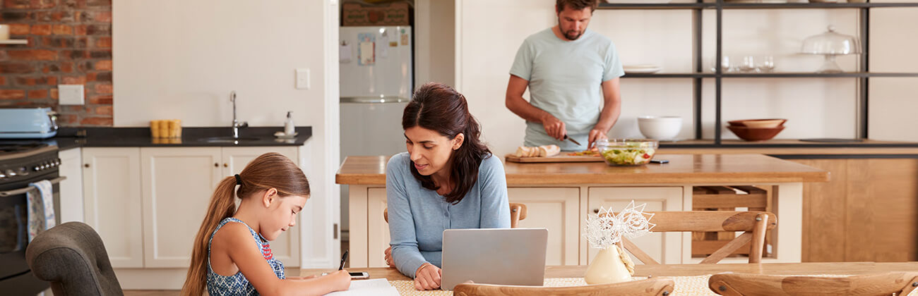 Mom on laptop helps daughter with homework at kitchen table while Dad fixes dinner