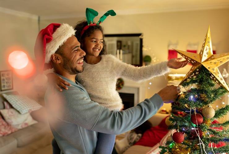 Daughter in holiday attire adds the star at the top of the Christmas tree with Dad holding her up to reach.