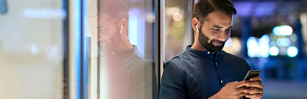 man leaning against window with earpods in and smiling at his phone at night